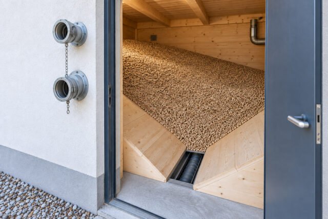 Geöffneter Pelletlagerraum mit Holzpellets, schrägem Holzboden und Befüllanschlüssen neben der Tür